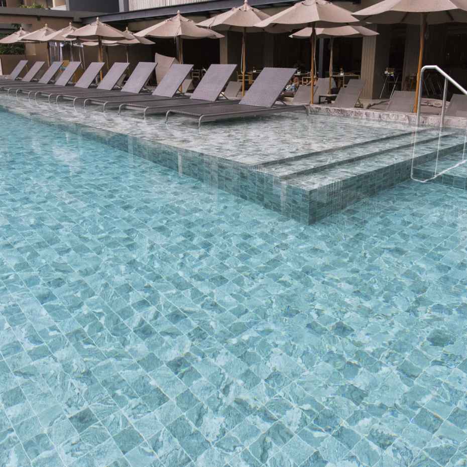A Swimming Pool Made of Alpine Beige 100x100 Mosaic Pool Tile at a Resort, with Pool Chairs Placed Next To It