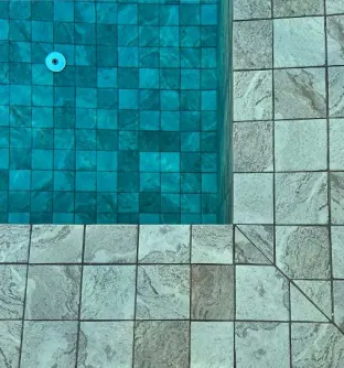 Textured deck tiles bordering a clear swimming pool lined with blue ceramic mosaic pool tiles.